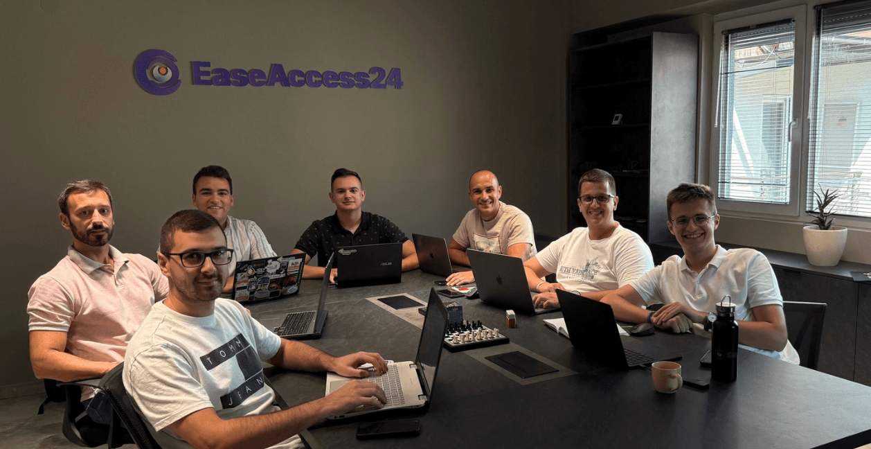 A team of seven people working at a large conference table with laptops in a modern office, with the EaseAccess24 logo visible on the wall behind them.