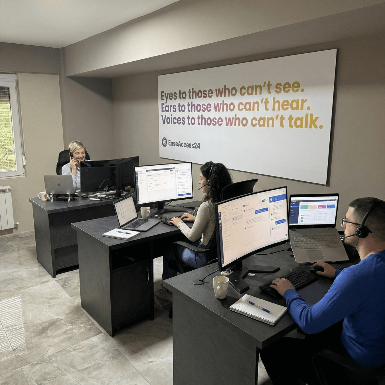 Three customer support agents wearing headsets work at desks with computers in a modern office. A large wall sign behind them reads, ‘Eyes to those who can’t see. Ears to those who can’t hear. Voices to those who can’t talk.’