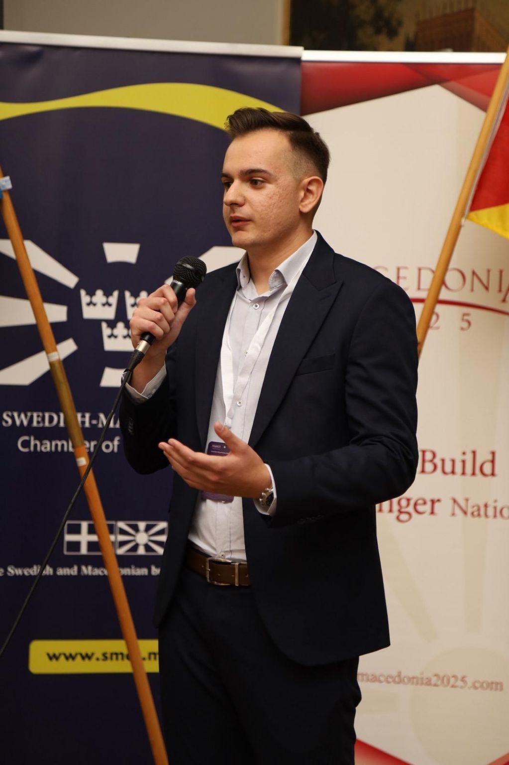 Filip Najdovski speaking with a microphone in front of SMCC and Macedonia2025 banners in Stockholm.